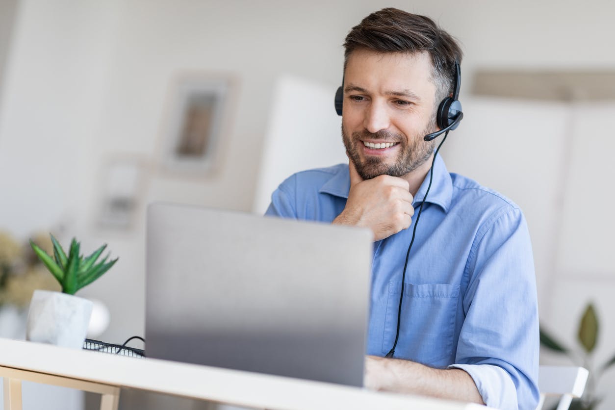 Homem usando um headset sorri com a mão no queixo olhando para o computador.
